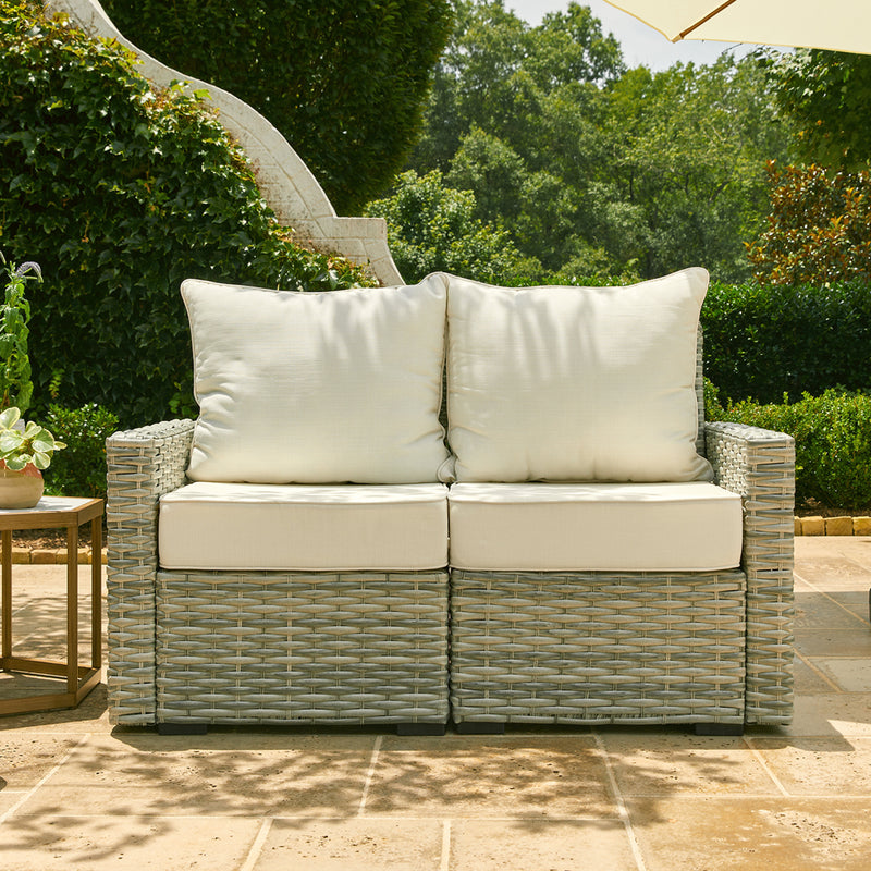 Gray Carmel Wicker Seat loveseat with beige cushions sits outdoors in front of a leafy greenscape. The edge of a table and an umbrella can be seen on the sides of the image.