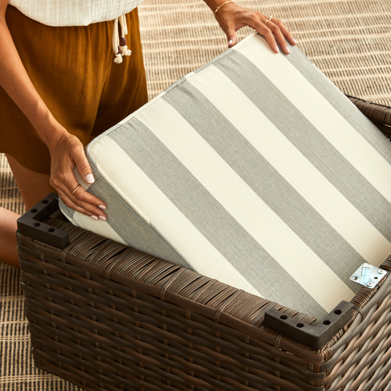 Top-down shot of the Carmel Wicker Seat, facing upside down. Hand on the left side of the image demonstrates the storage features, tucking cushions into the empty underside of the chair.